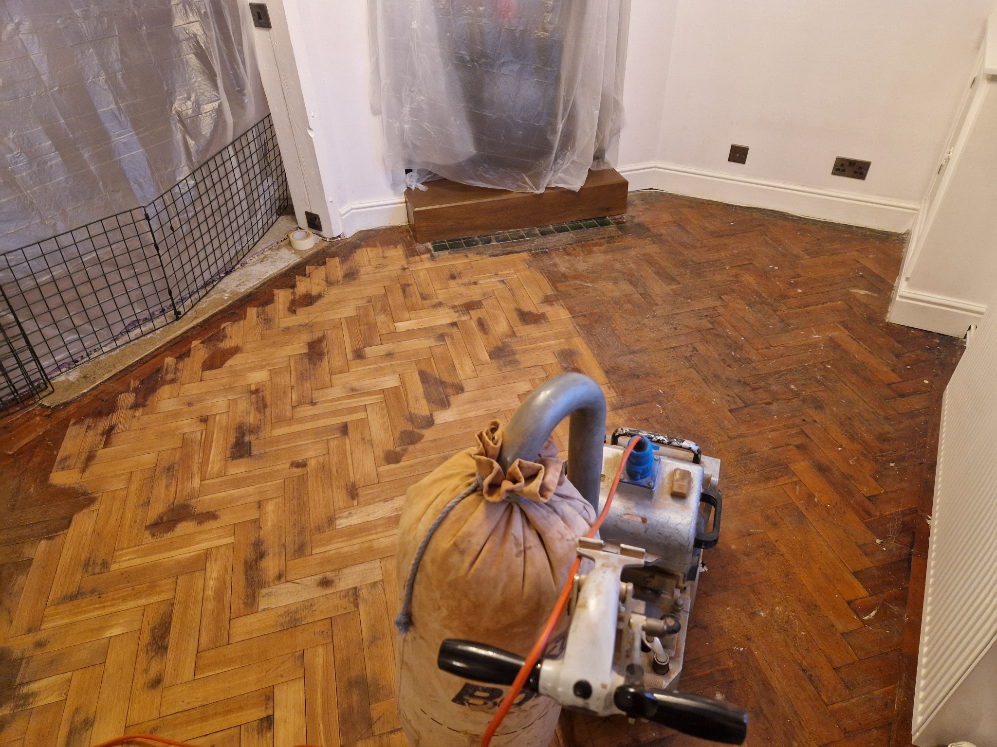 A partially sanded herringbone oak parquet floor with a drum sander in the foreground, illustrating the wood floor sanding process in progress.