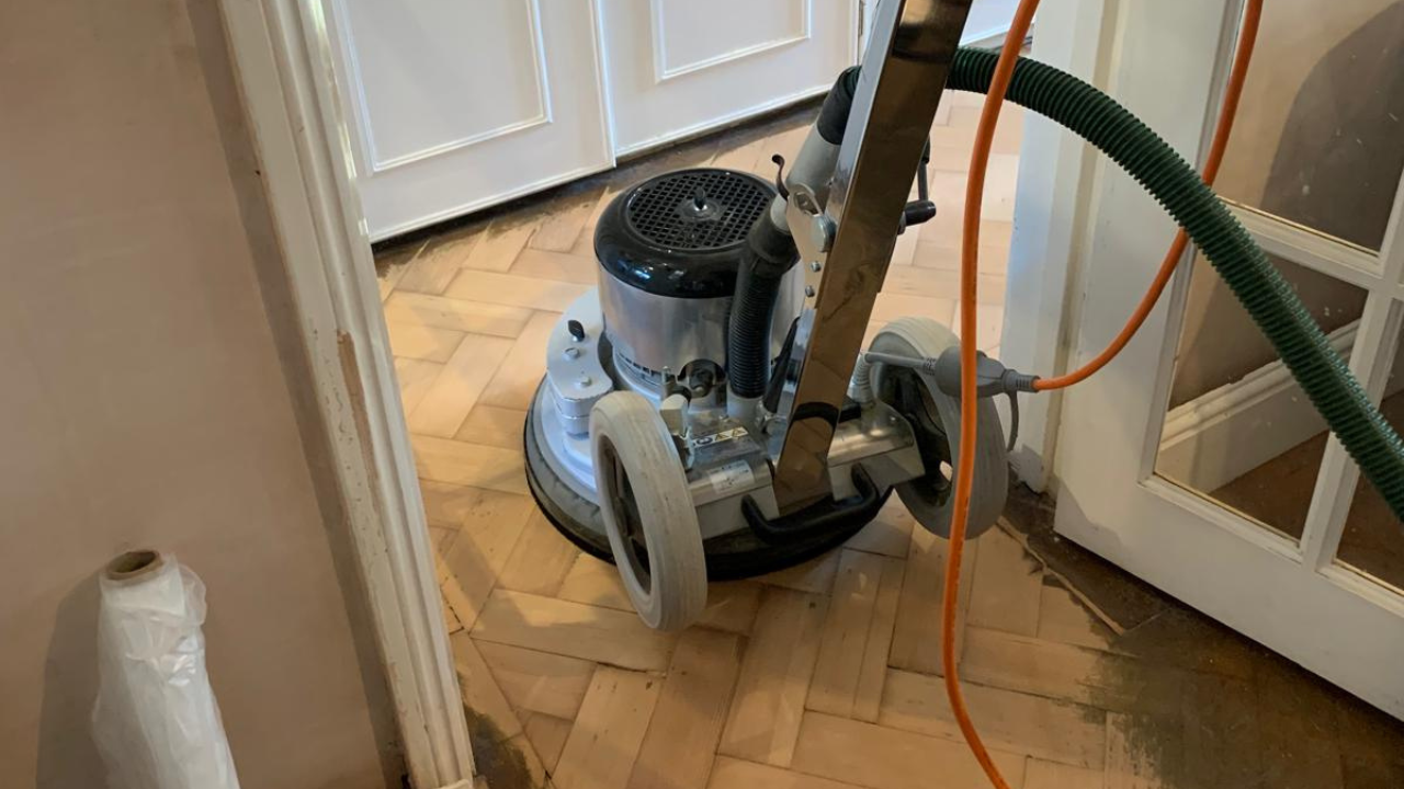 A specialized floor sanding machine operating on a herringbone oak parquet floor, removing the old finish and preparing the surface for refinishing.