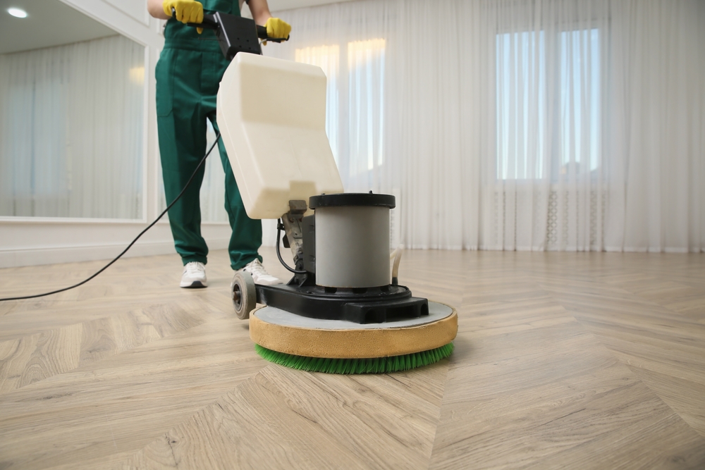 A technician in green workwear operating a floor buffer on a chevron-patterned oak floor, emphasizing wood floor sanding and refinishing.