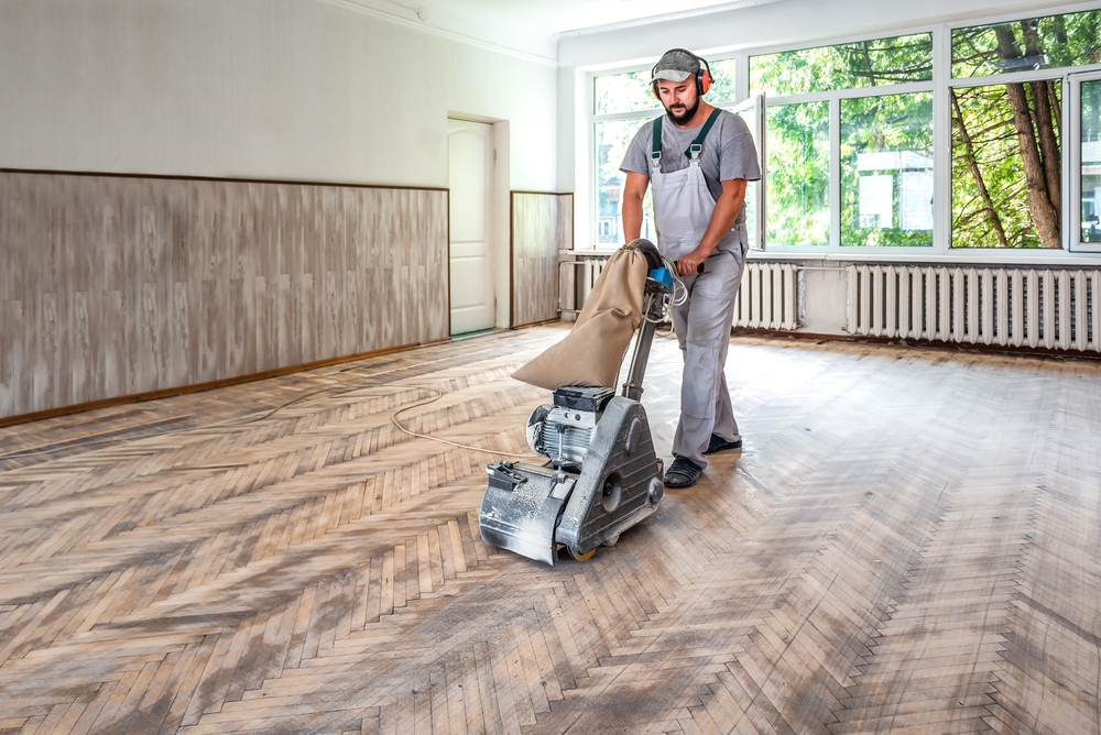 A flooring specialist using a drum sander to refinish a herringbone oak parquet floor, highlighting a wood floor sanding process in a large, light-filled room.