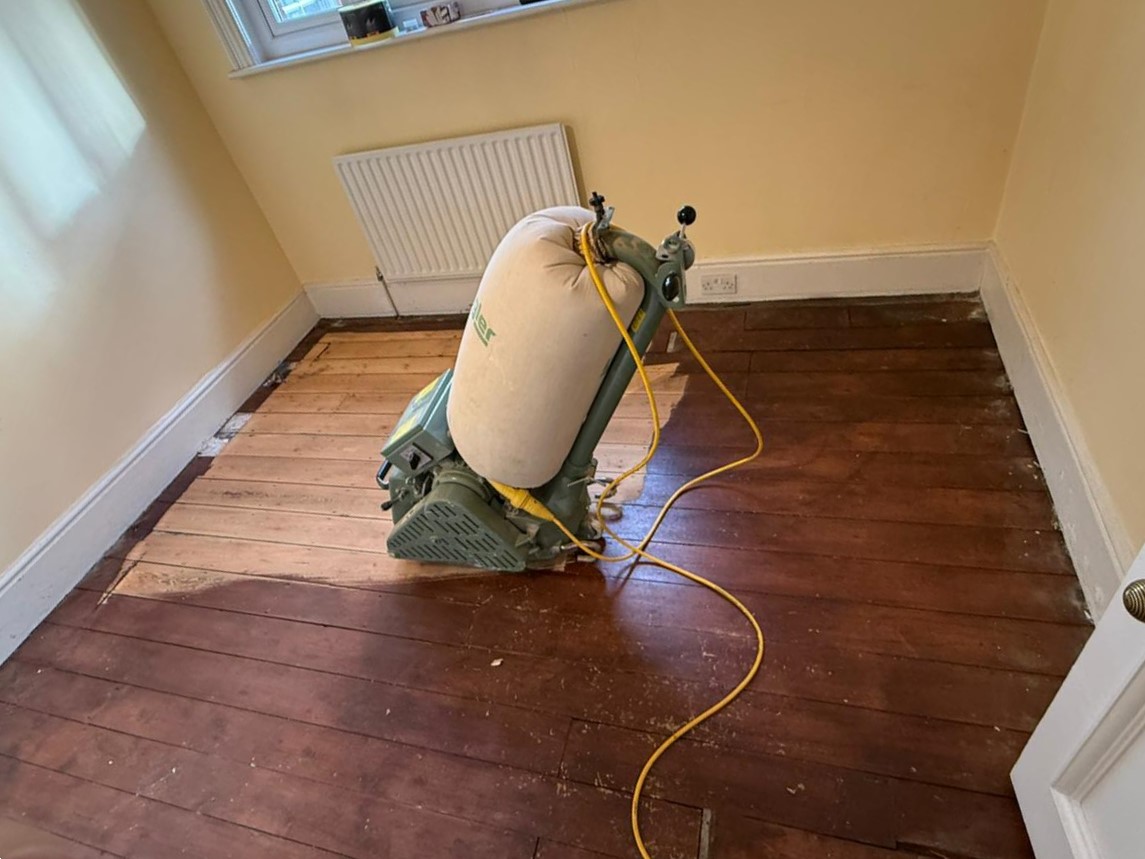 Wood Floor Sanding in West Brompton bedroom on pine softwood floorboards, showing part-sanded boards beside dark old finish with a drum sander.