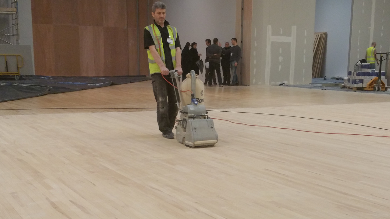 Wood Floor Sanding in a West Brompton sports hall, with a contractor sanding a maple hardwood floor using a professional belt sander.