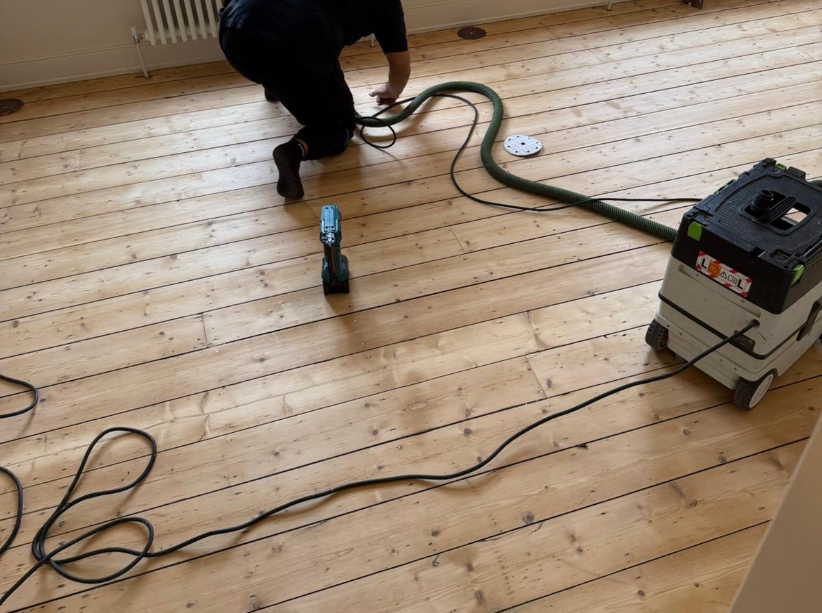 Wood Floor Sanding in West Brompton on pine softwood floorboards, contractor working on hands and knees with dust-extraction vacuum.