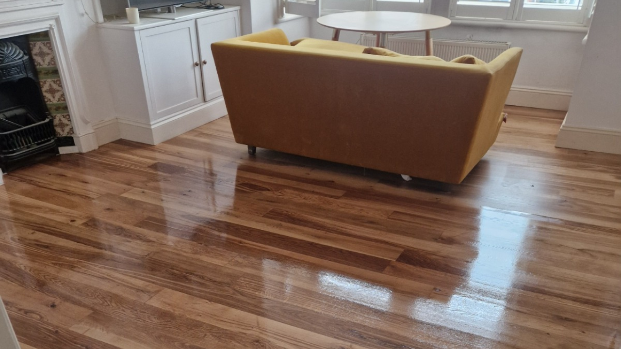 Hardwood Floor Sanding in West Brompton on an oak plank hardwood living room floor, shown freshly refinished to a high gloss around a mustard sofa and fireplace.
